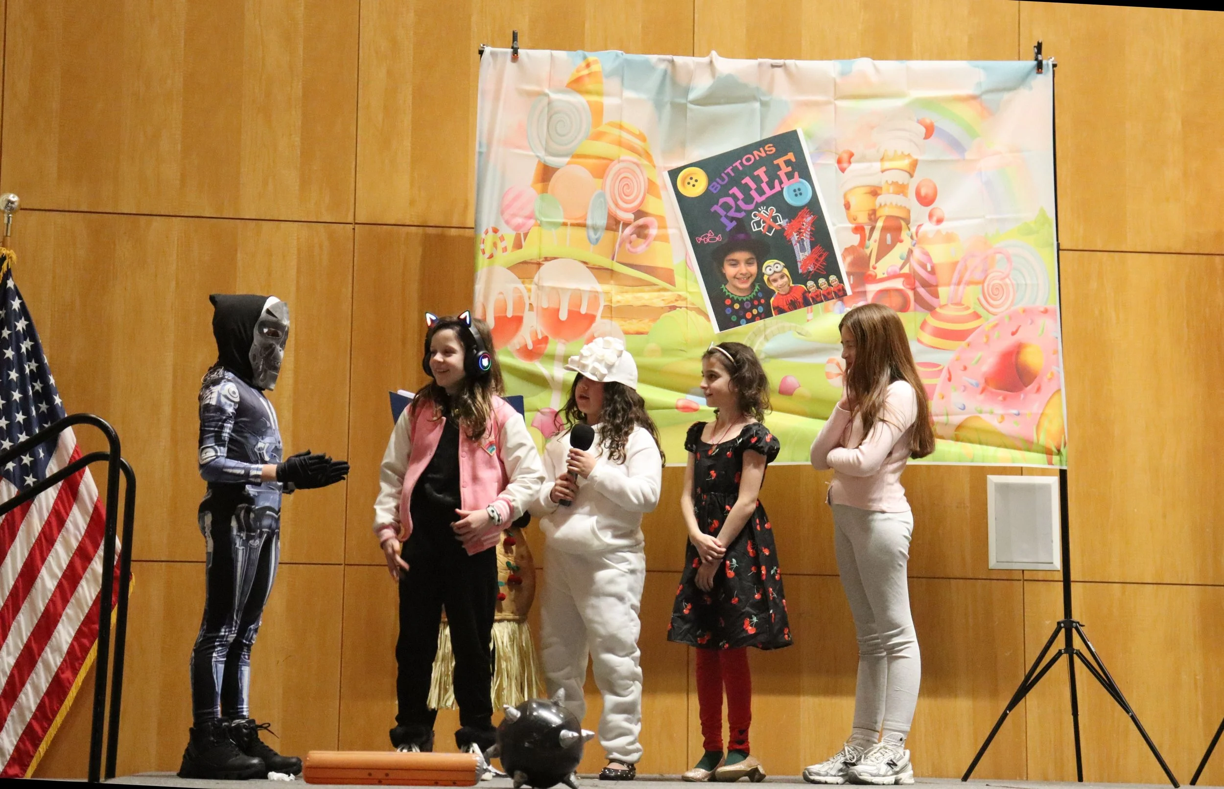 Children dressed in costumes standing on a stage, participating in a performance or event, with a colorful backdrop featuring candy and sweets.