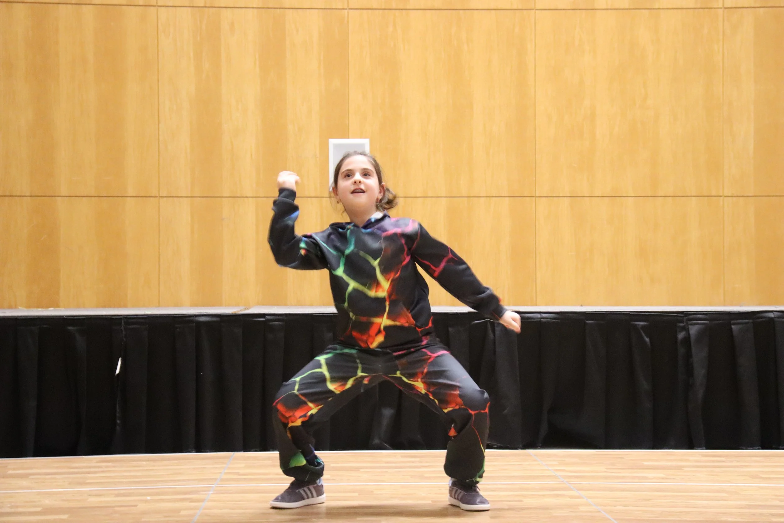 A young girl dancing on a stage with a wooden wall background, wearing a black outfit with a colorful lightning pattern, mid-move with one arm raised and smiling.