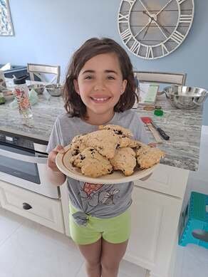 A young girl with shoulder-length brown hair smiling and holding a large plate of cookies in a kitchen.