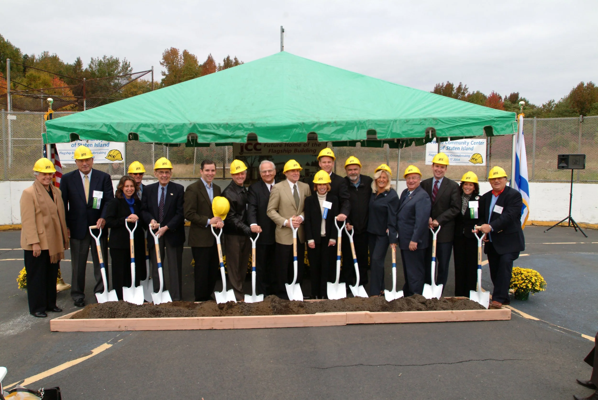 Group of people wearing yellow construction helmets standing in front of a green tent and holding shovels during a groundbreaking ceremony.