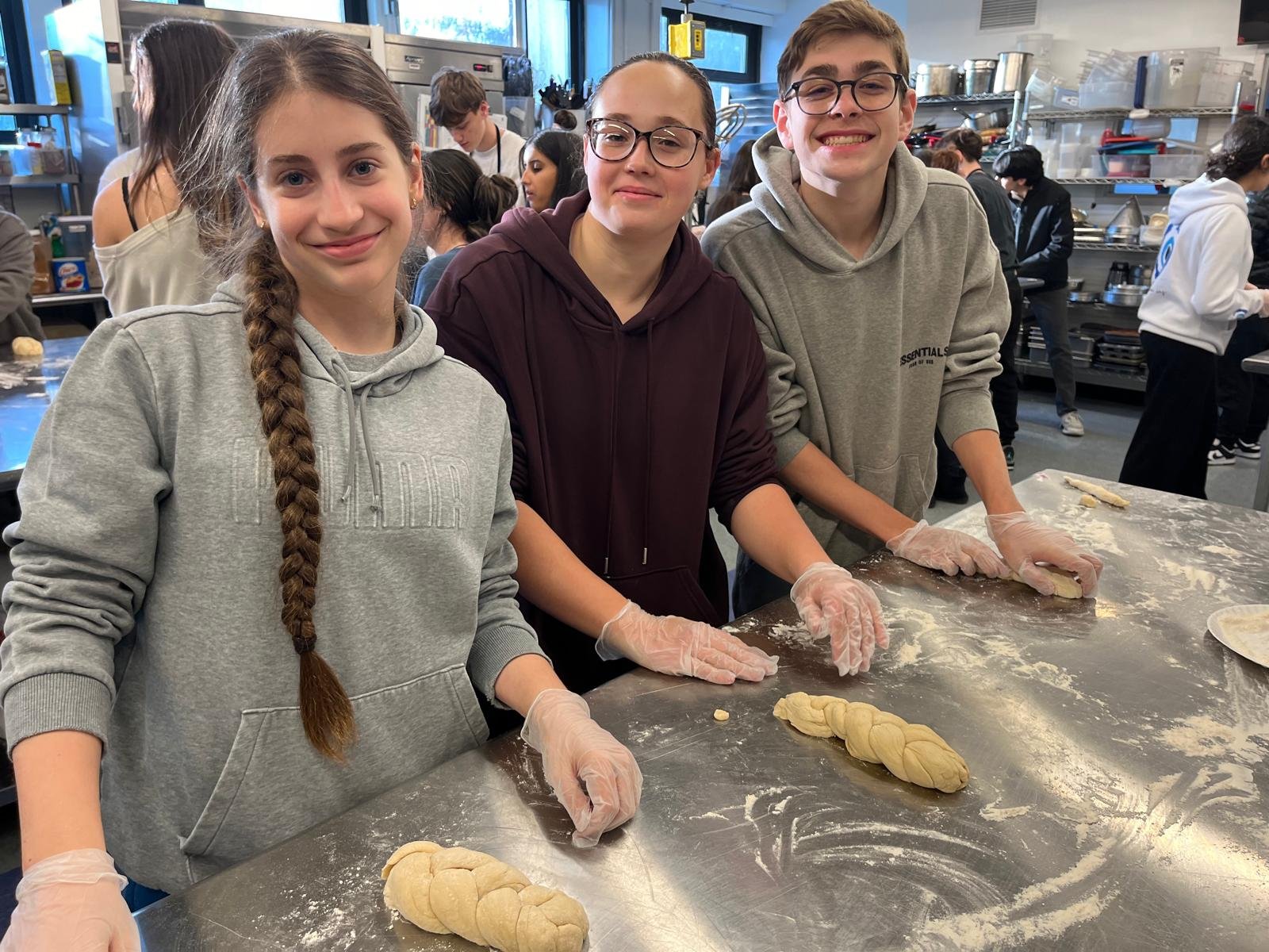 Three teenagers wearing gloves making braided dough in a commercial kitchen with other people working in the background.