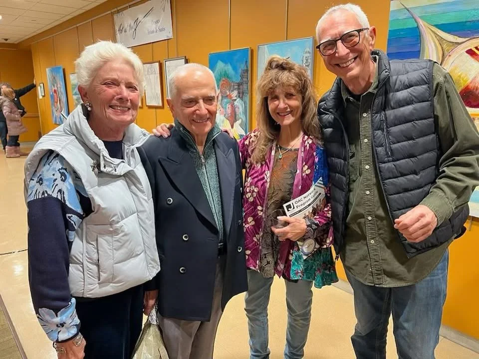 Four people smiling and posing for a photo at an art gallery with paintings on the wall behind them.