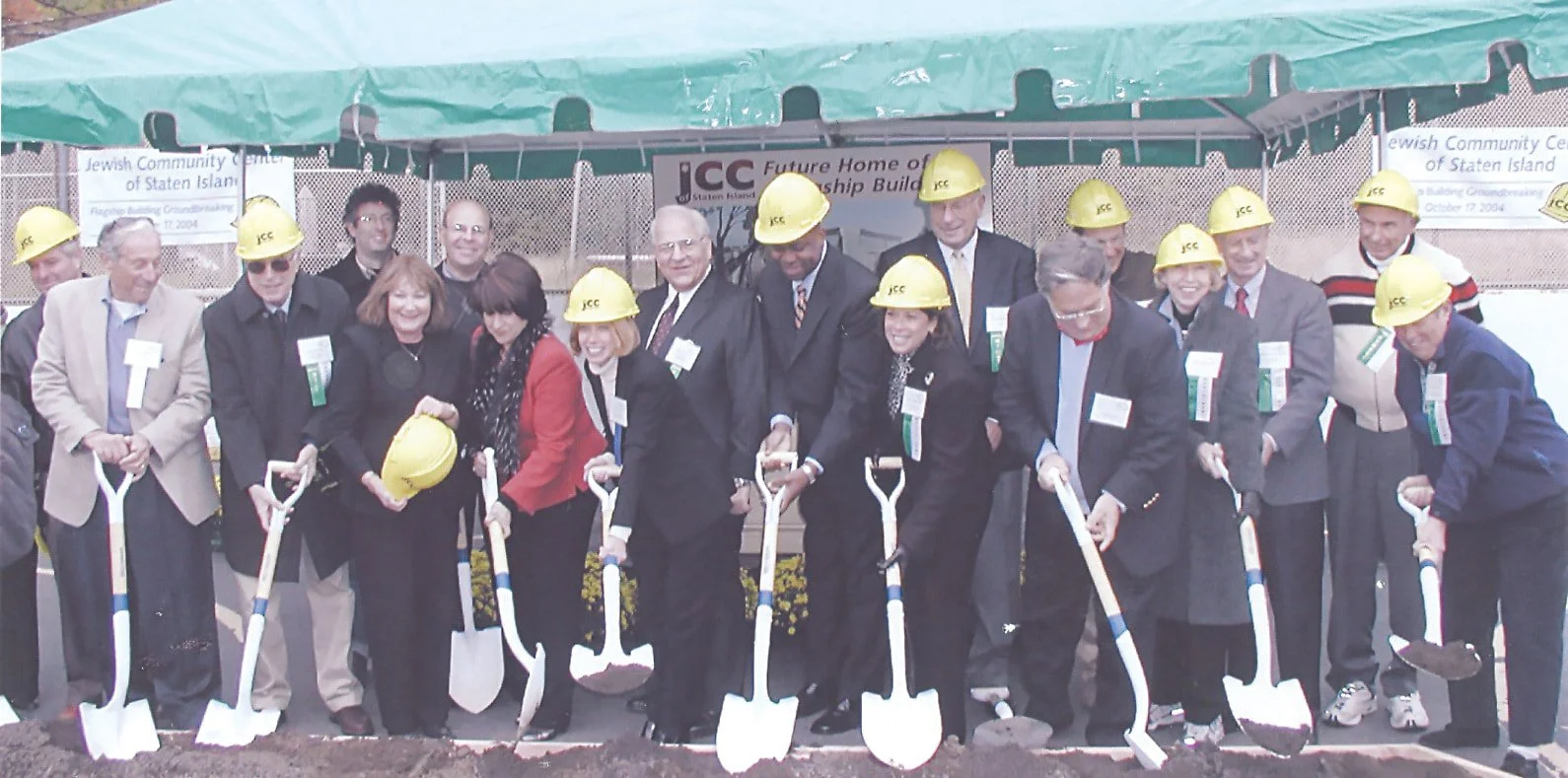 Group of people at a groundbreaking ceremony, wearing yellow construction helmets and formal business attire, holding shovels.