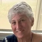 Close-up of a smiling woman with short, gray hair, wearing a blue top, indoors with a light-colored wall behind her.
