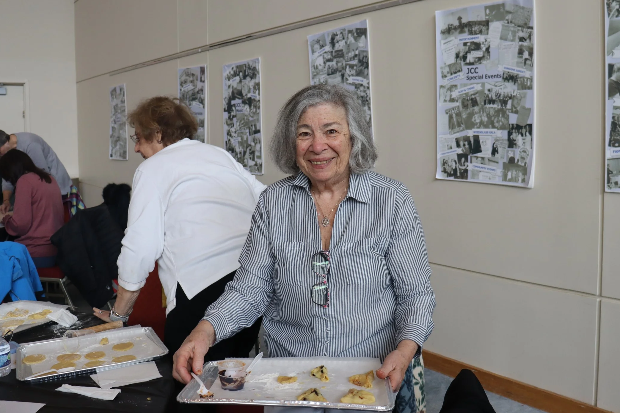An elderly woman with gray hair smiling at the camera, wearing a striped shirt, standing at a table with food. In the background, several people are gathering in a room decorated with collage posters on the wall.