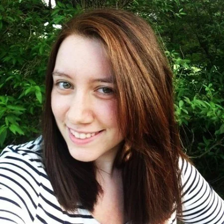 Young woman with shoulder-length red hair smiling outdoors, wearing a striped shirt, surrounded by green foliage.