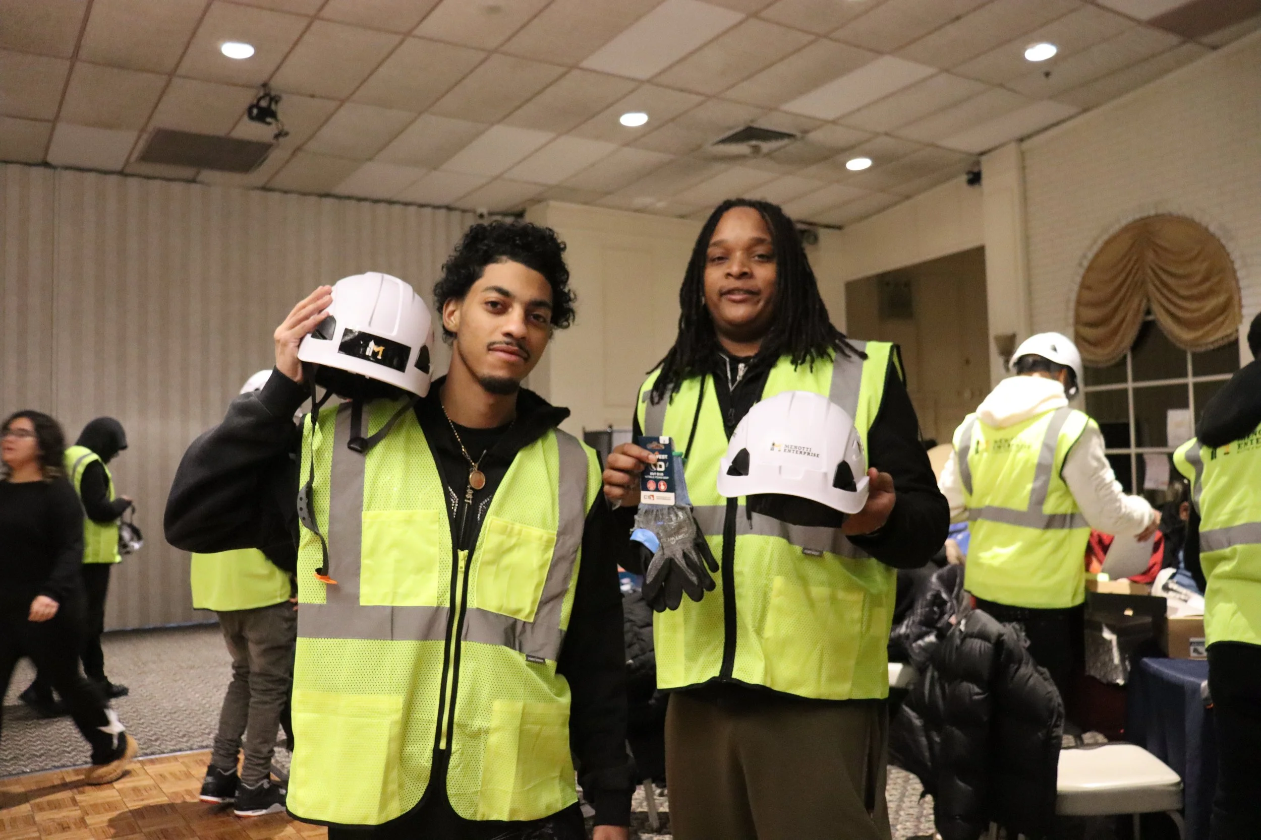 Two young men in high-visibility safety vests holding helmets and showing ID badges at an indoor event, with other people in safety gear in the background.