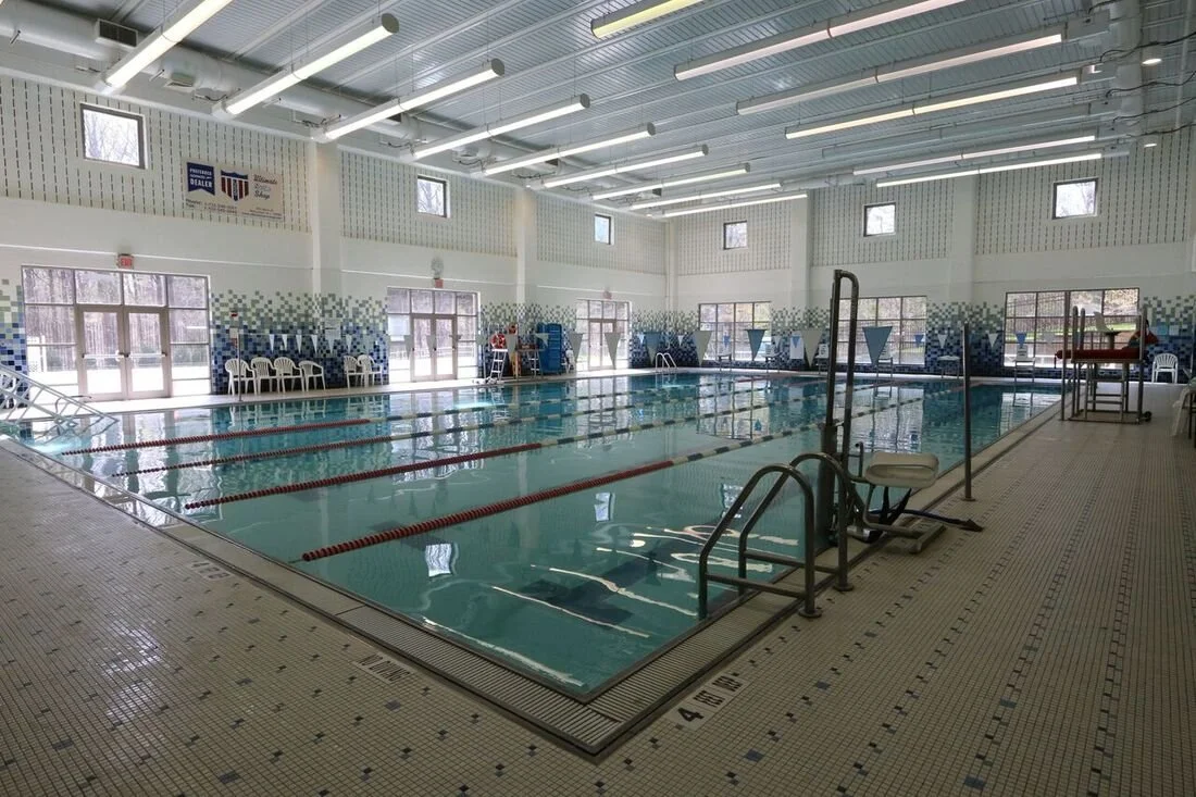 Indoor swimming pool with lanes, chairs, and windows, illuminated by overhead lights.
