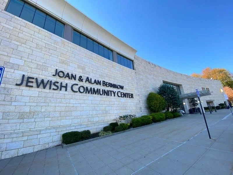 Exterior view of the Joan & Alan Bernick Jewish Community Center building with a stone facade, windows, trees, bushes, and a sidewalk in front.