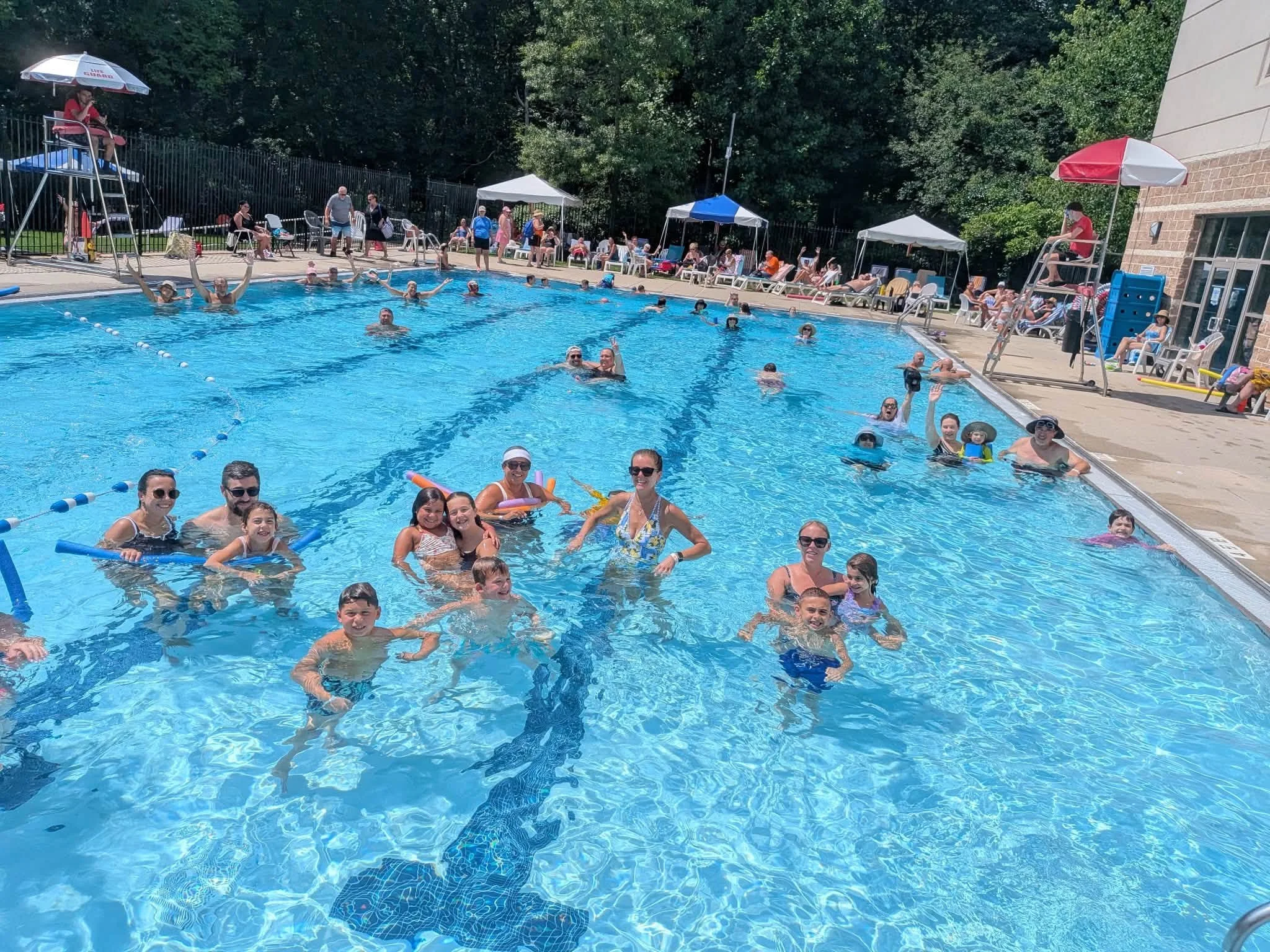 People swimming, playing, and relaxing at an outdoor swimming pool area on a sunny day, with poolside chairs, umbrellas, and a building in the background.