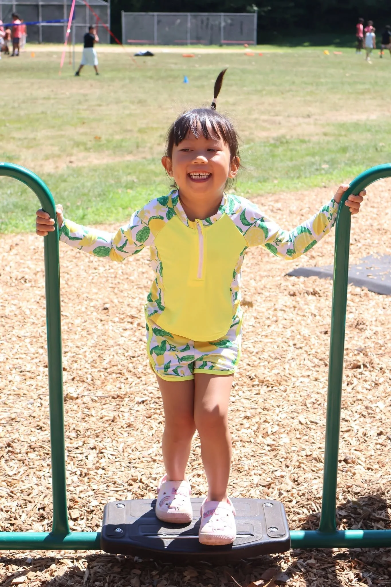 A young girl smiling and standing on a playground step platform. She is holding onto green bars and wearing a yellow and green lemon-patterned outfit with pink shoes. Other children are playing in the background on a sunny day.