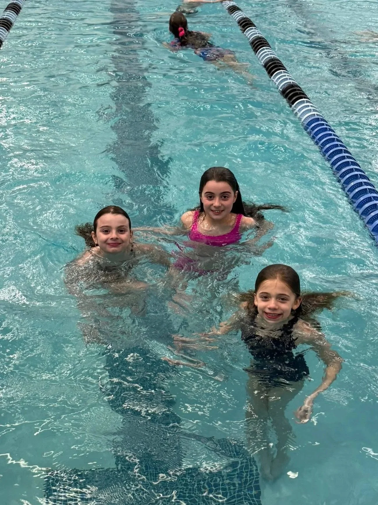 Four young girls swimming in a pool, smiling at the camera. One girl is in a black swimsuit, another in a pink swimsuit, another in a black swimsuit with a pattern, and the fourth is in a black swimsuit. The swimming pool has blue tiles and lane dividers.