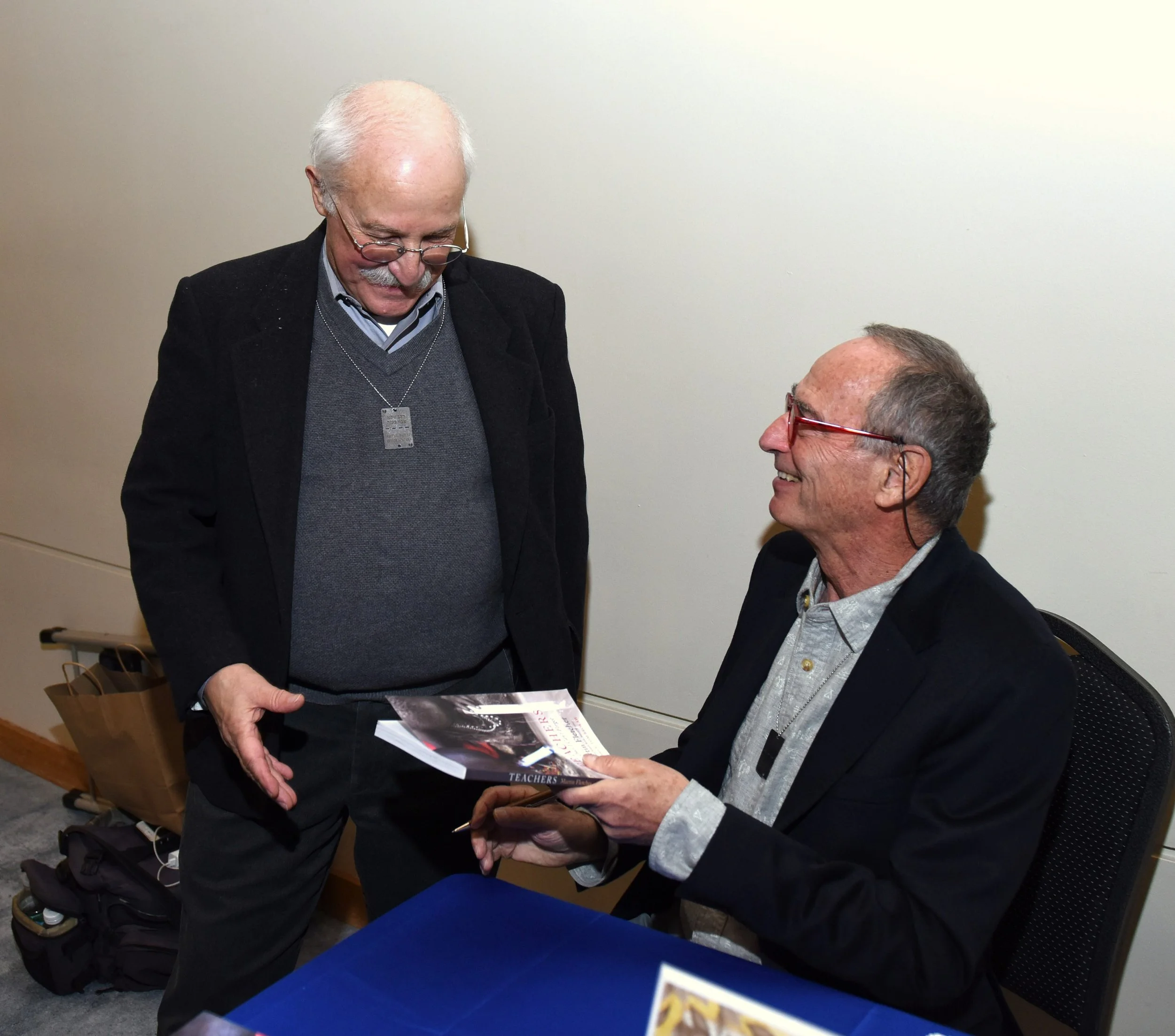 Two elderly men engaged in a conversation, one standing and the other sitting. The man standing has white hair, glasses, a dark blazer, and is holding a booklet or magazine. The man sitting has gray hair, glasses, and is smiling while also holding the booklet. They are in an indoor setting with a plain light-colored wall in the background.