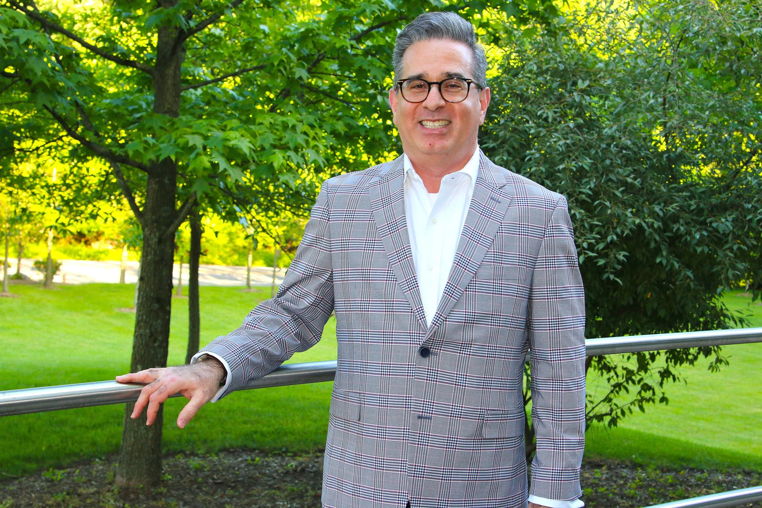 A smiling man with glasses, wearing a light-colored plaid blazer and white shirt, leaning on a metal railing outdoors with trees and grass in the background.