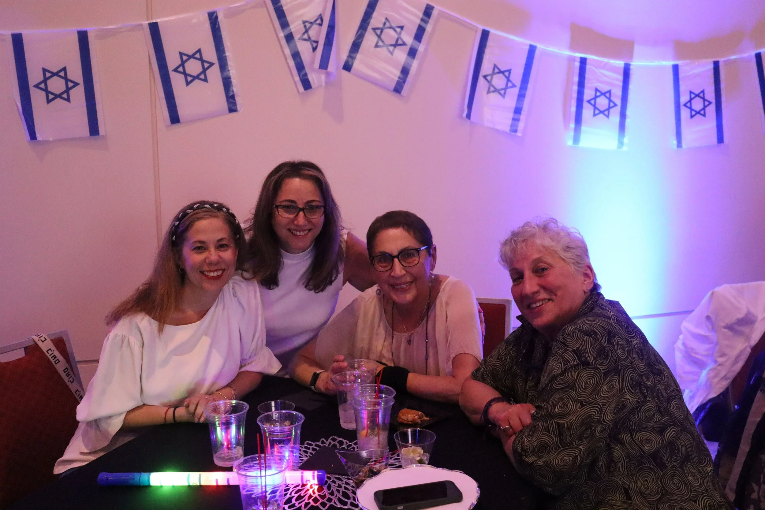 Group of five women sitting around a table at an indoor party with Israeli flags hanging from the ceiling.