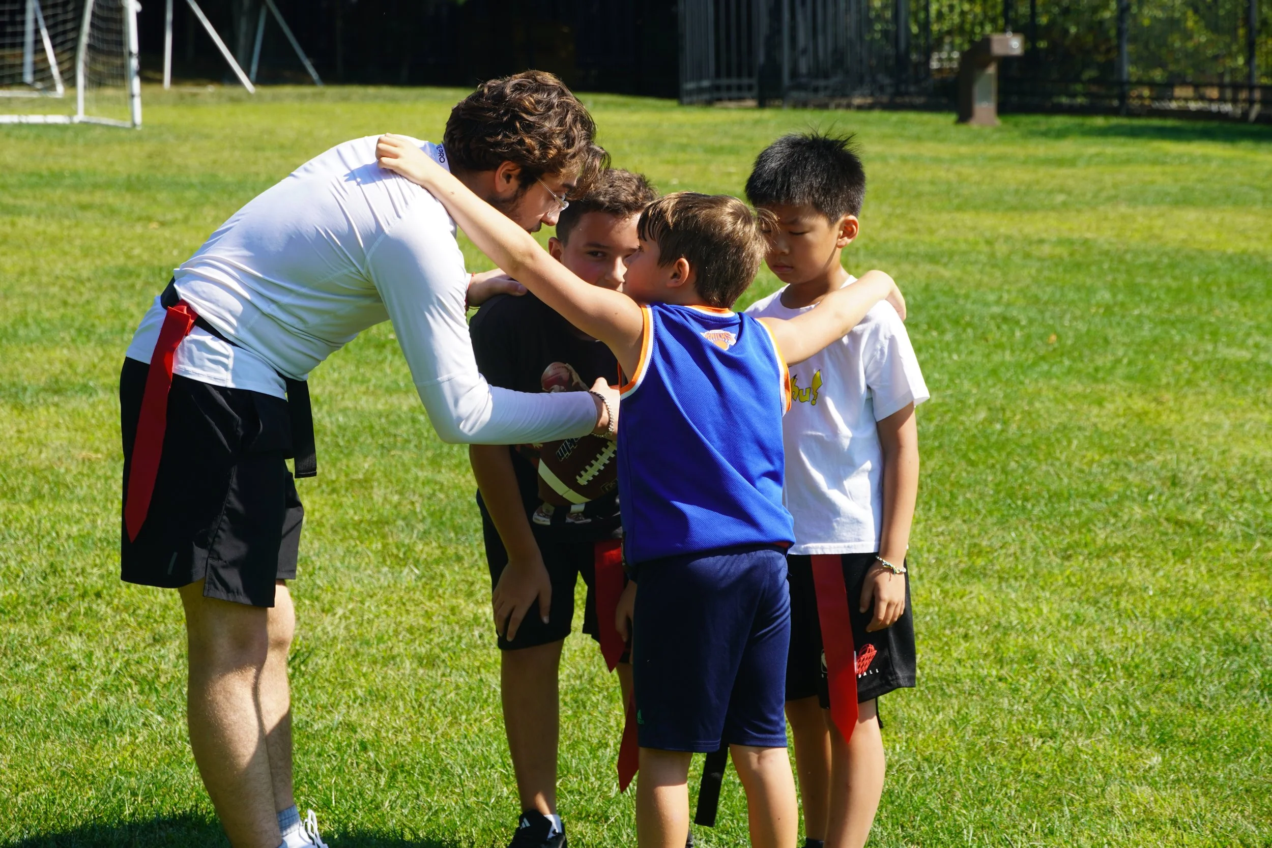A group of young boys standing in a line with their arms around each other, receiving instructions from a coach on a grassy field during daytime.
