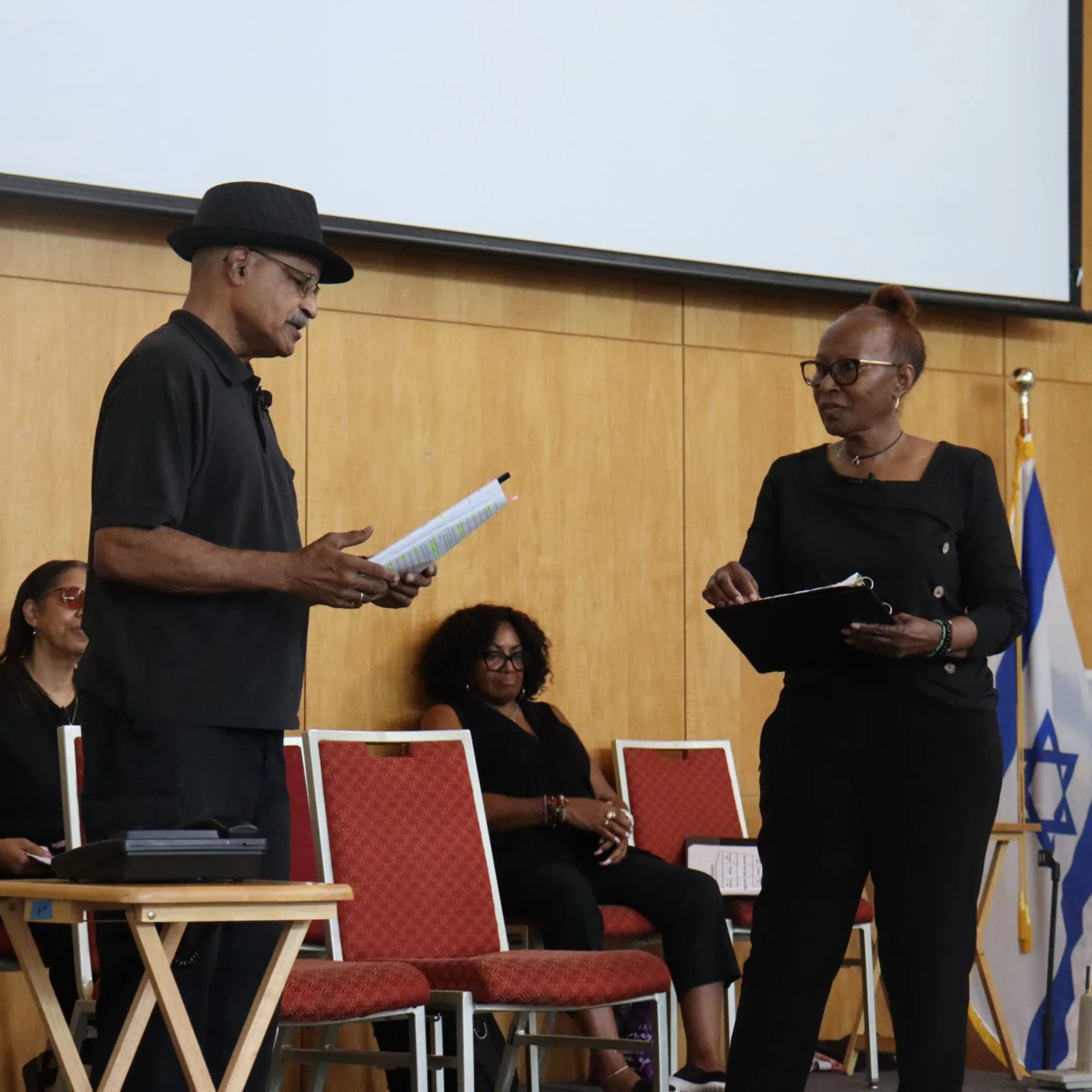 A man wearing a black hat, black shirt, and glasses stands on the left, reading from a paper, while a woman in black attire and glasses stands on the right holding a folder, engaging in conversation during a formal event with seated women and an Israeli flag in the background.
