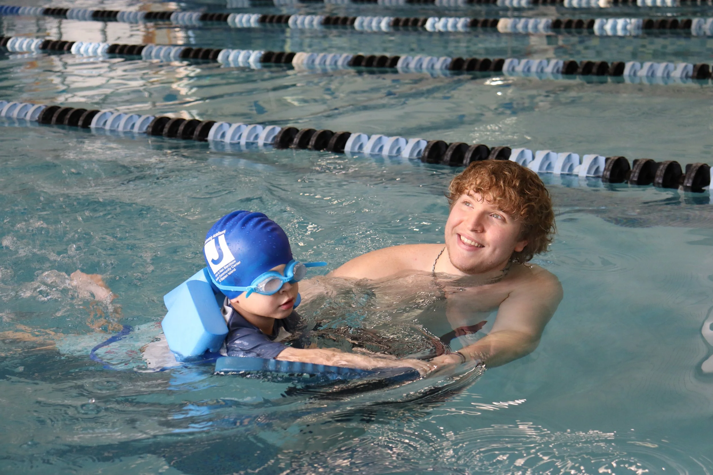 A young man with curly hair in a swimming pool helping a young child with a blue swim cap and goggles learn to swim.
