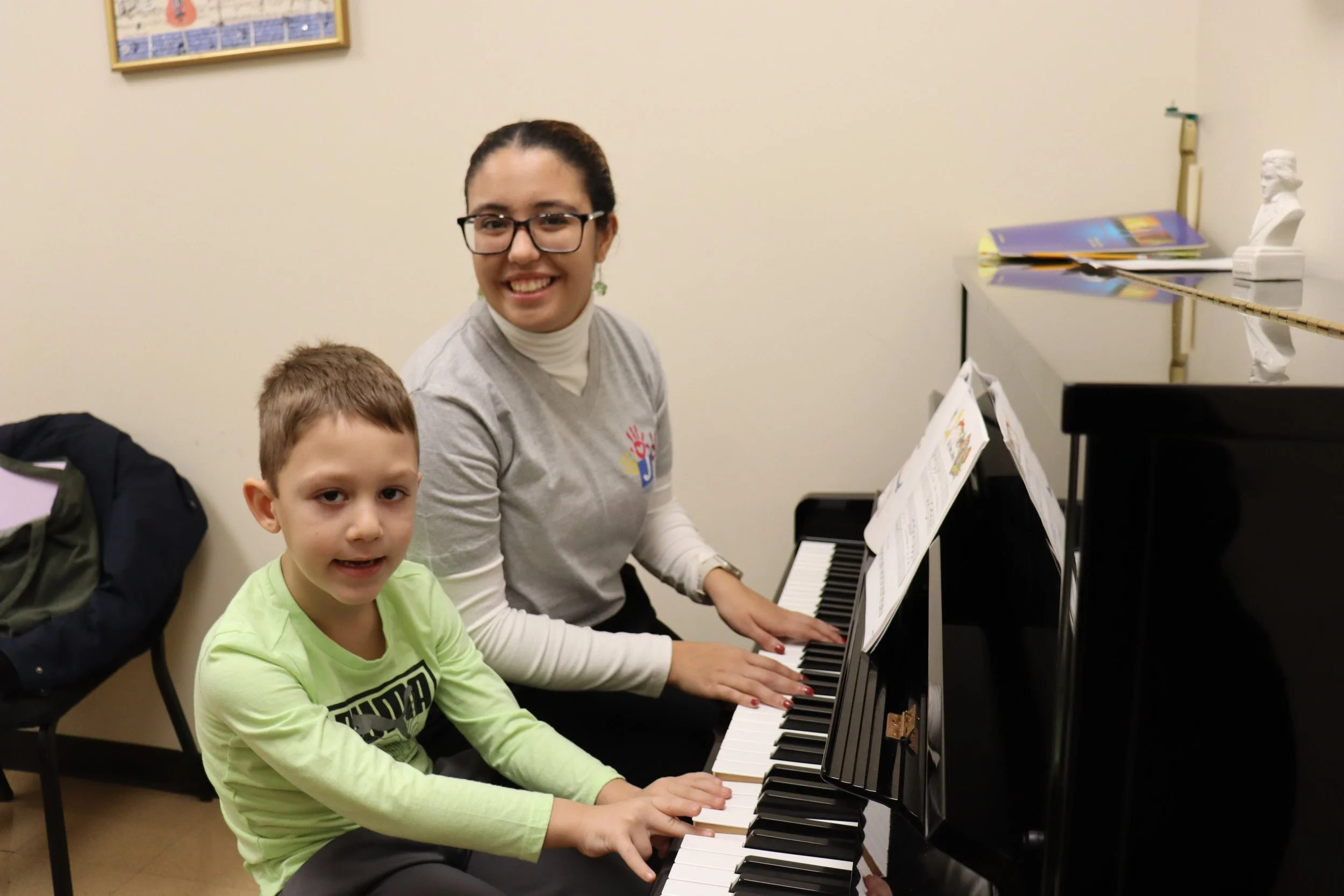 A woman and a young boy playing the piano together in a music room, with music sheets on the stand and colorful books on a table nearby.