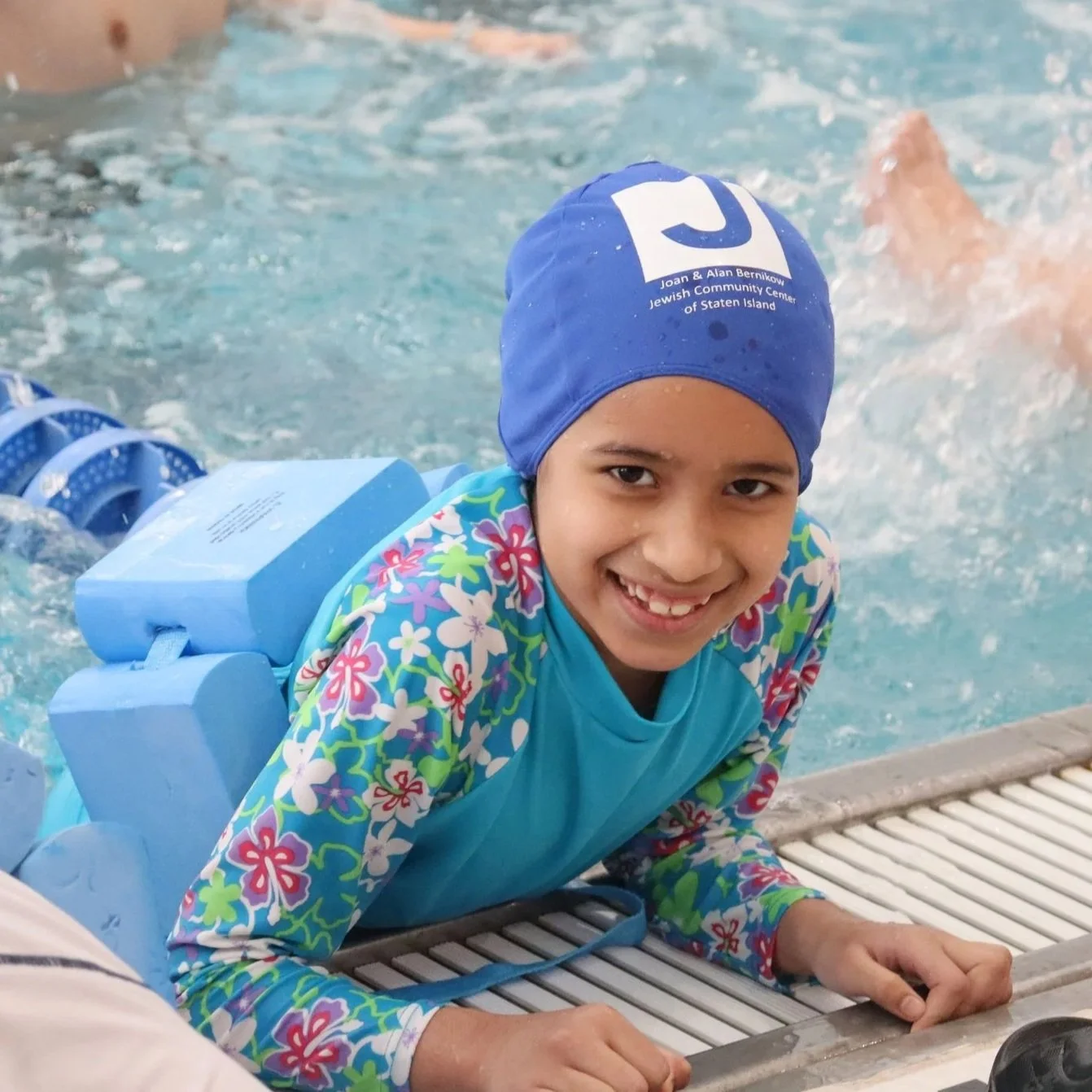 A young girl in a colorful floral swimsuit and blue swim cap smiling by the poolside, with swim training equipment attached to her back.