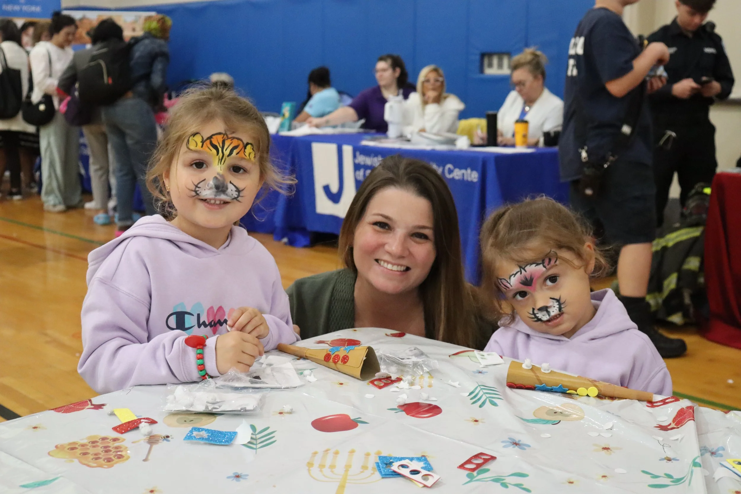 Two young girls with face paint of animals and a woman smiling, seated at a table with festive decorations, in a gymnasium with people and tables in the background.