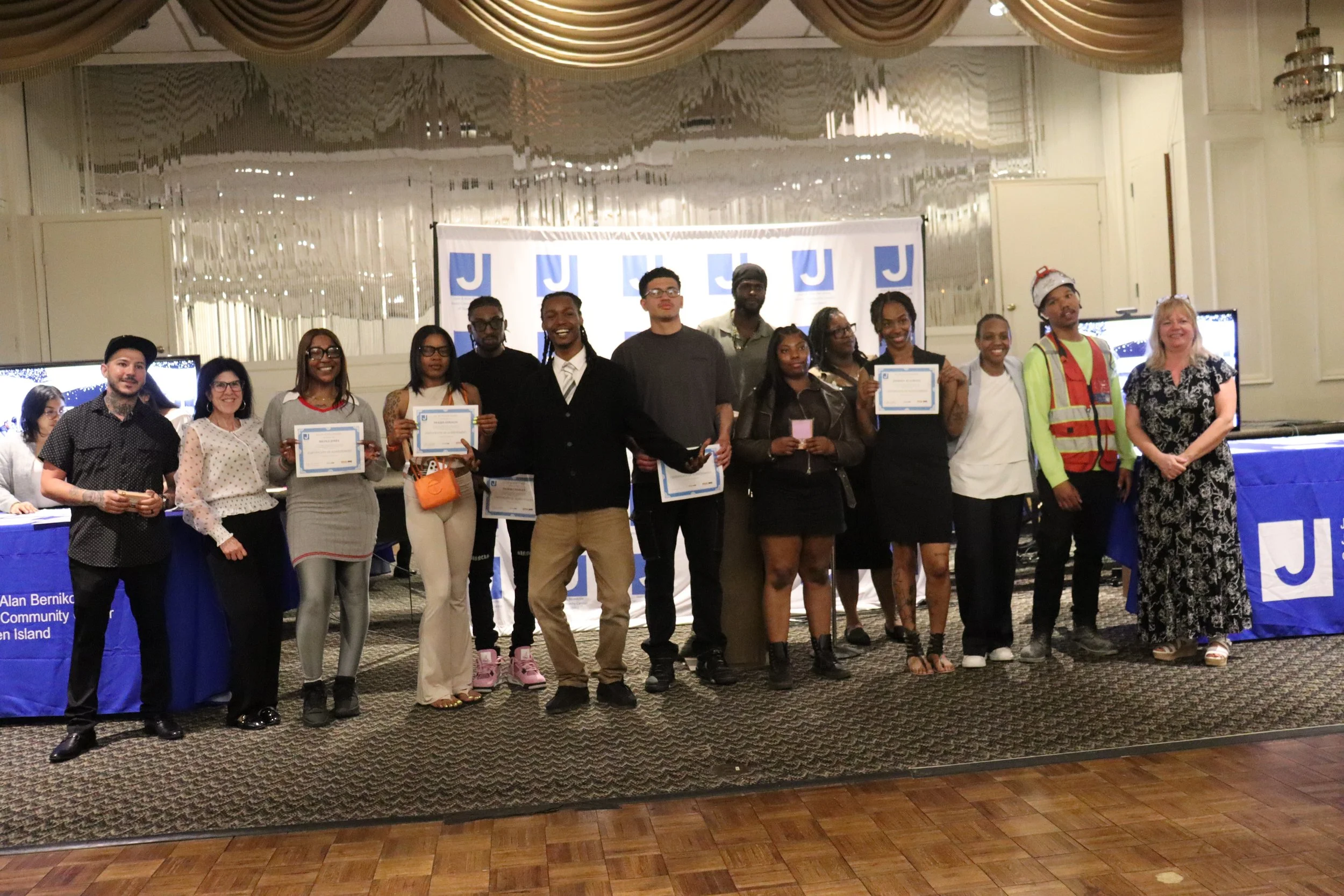 A diverse group of men and women posing with certificates at an indoor event, with a backdrop displaying the letter J.