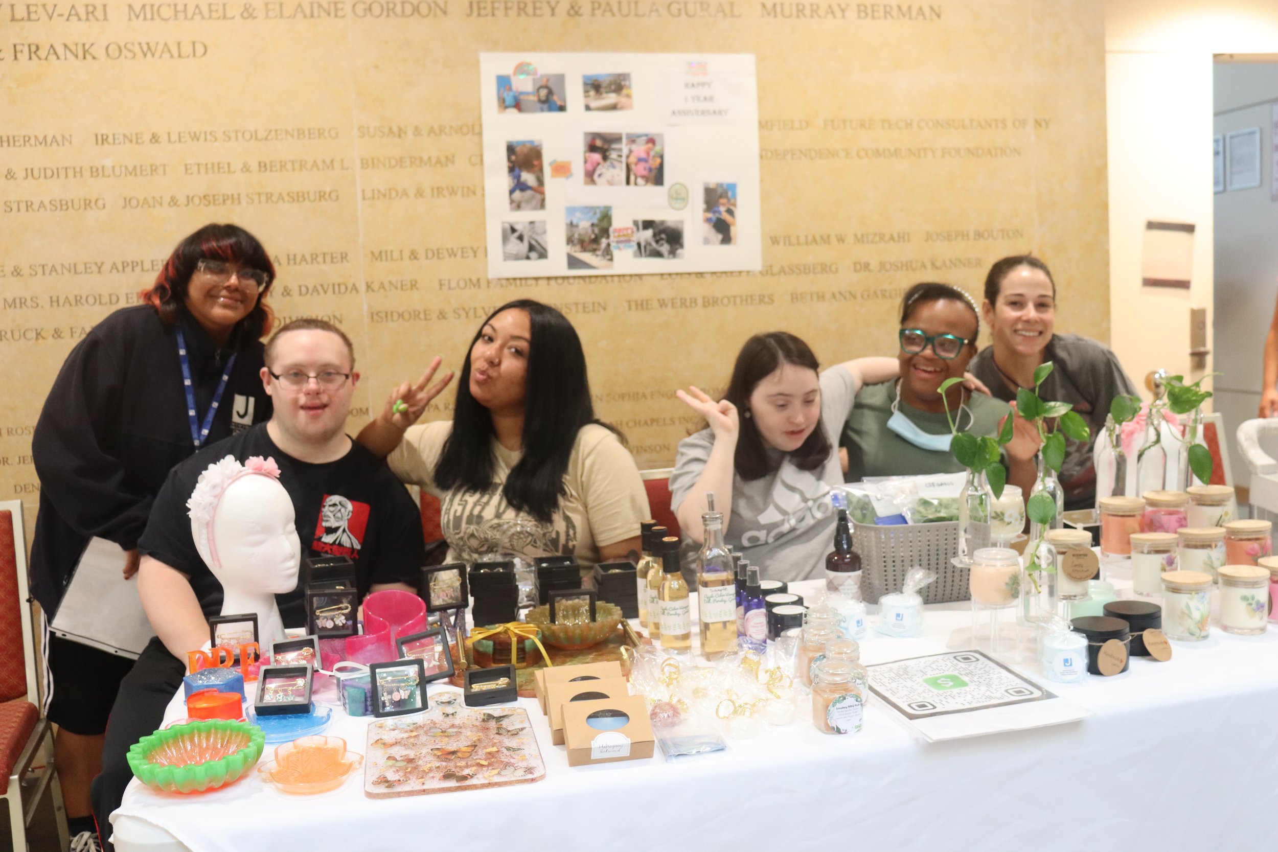 Group of five women and one young man are sitting at a table displaying various jewelry and beauty products. The women are smiling and making peace signs. The background has a wall with text and a poster with photos, and the table is decorated with plants and containers of products.