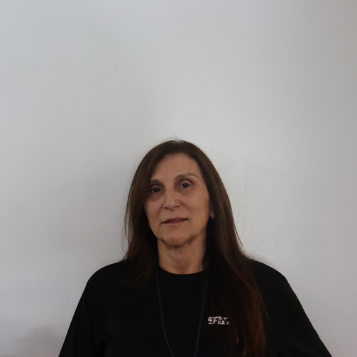 A woman with long brown hair wearing a black shirt standing against a plain white wall.