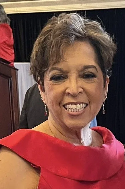 Close-up of a smiling woman with short brown hair wearing a red dress and earrings, at an indoor event.