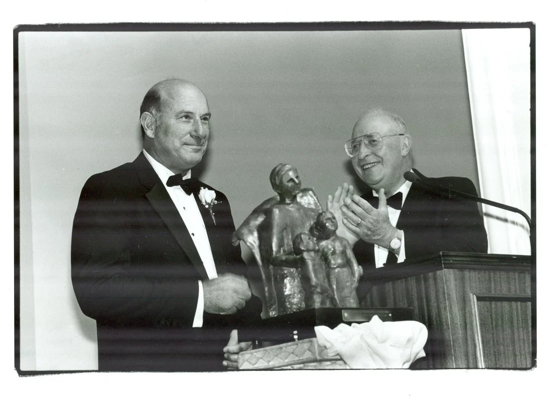 A black-and-white photo of two men in tuxedos at an award ceremony, with one man handing a religious sculpture to the other, both smiling.
