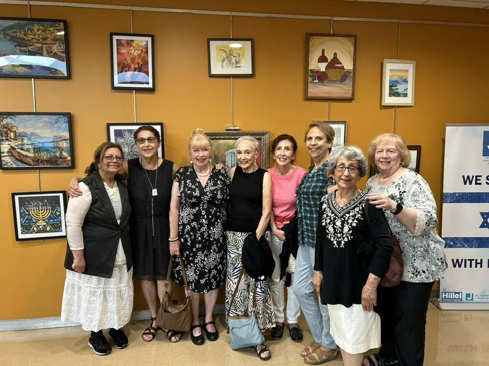 A group of nine women standing side by side indoors in front of a yellow wall decorated with framed artwork, smiling for the camera.