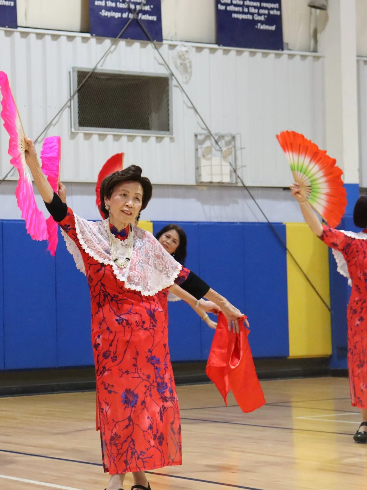 Women in red floral dresses performing a fan dance indoors, holding colorful fans and red cloths, with a blue and yellow gymnasium wall in the background.