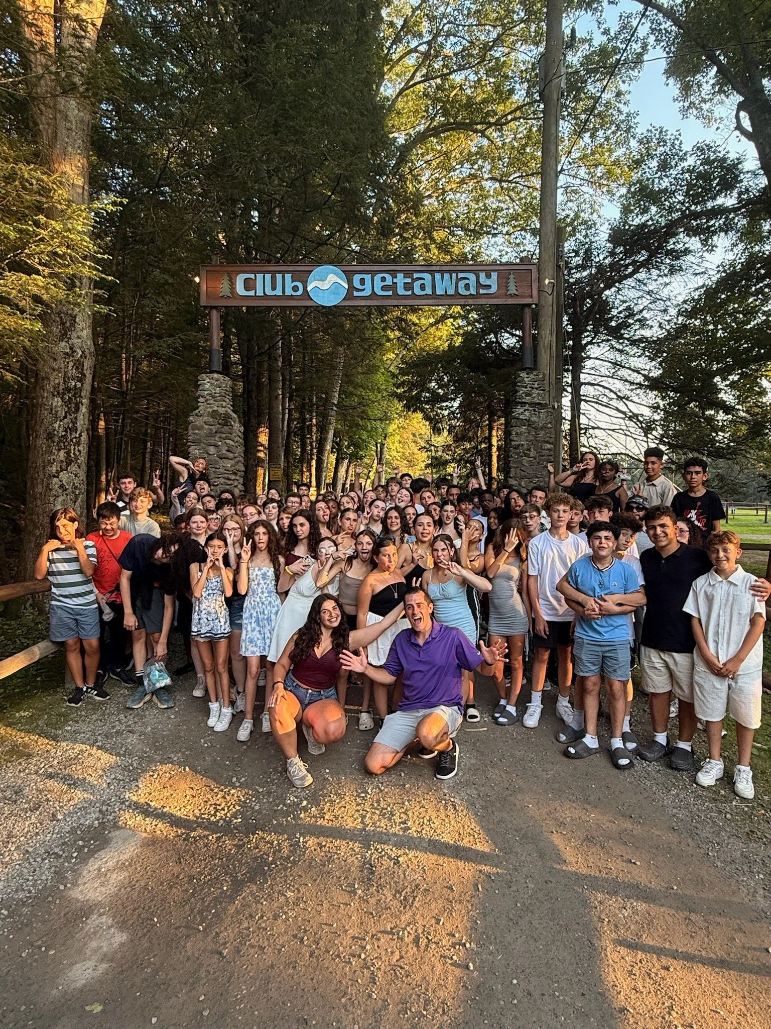 Large group of young people and some adults posing outdoors under a sign that reads 'Club Getaway' in a forested area.