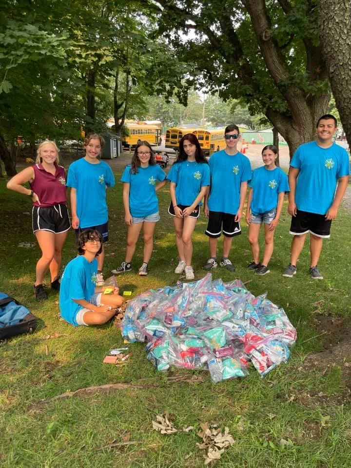 Group of seven children and teenagers standing behind a large pile of plastic water bottles on the grass in a park with trees, two yellow school buses, and a playground in the background.