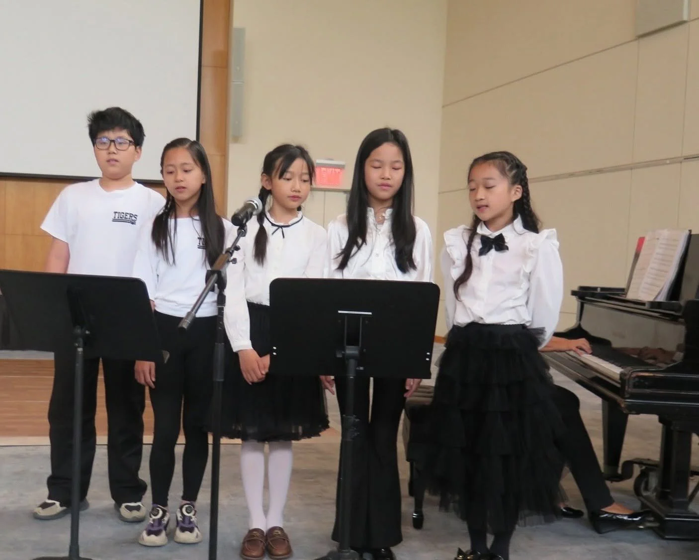 Five children singing or performing on stage, standing behind music stands in front of a microphone with a piano nearby.