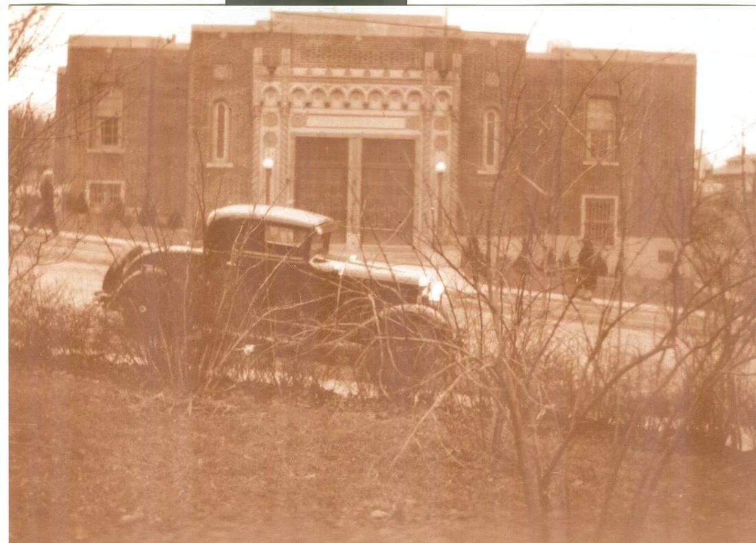 An old black and white photograph of a brick building with ornate architectural details and a large door, a vintage car parked in front, and some people walking on the sidewalk.
