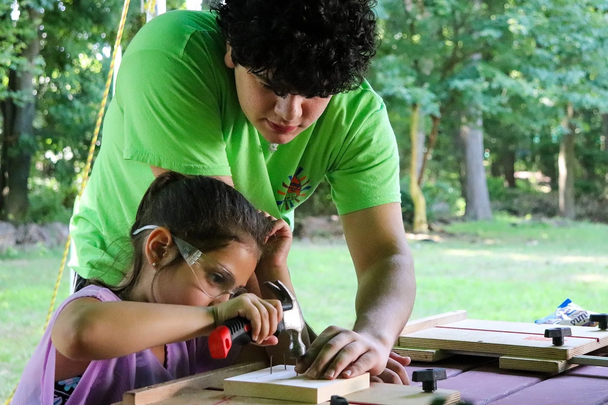 A young girl using a drill to work on a piece of wood at an outdoor woodworking project. An adult, possibly a counselor or teacher, is assisting her.