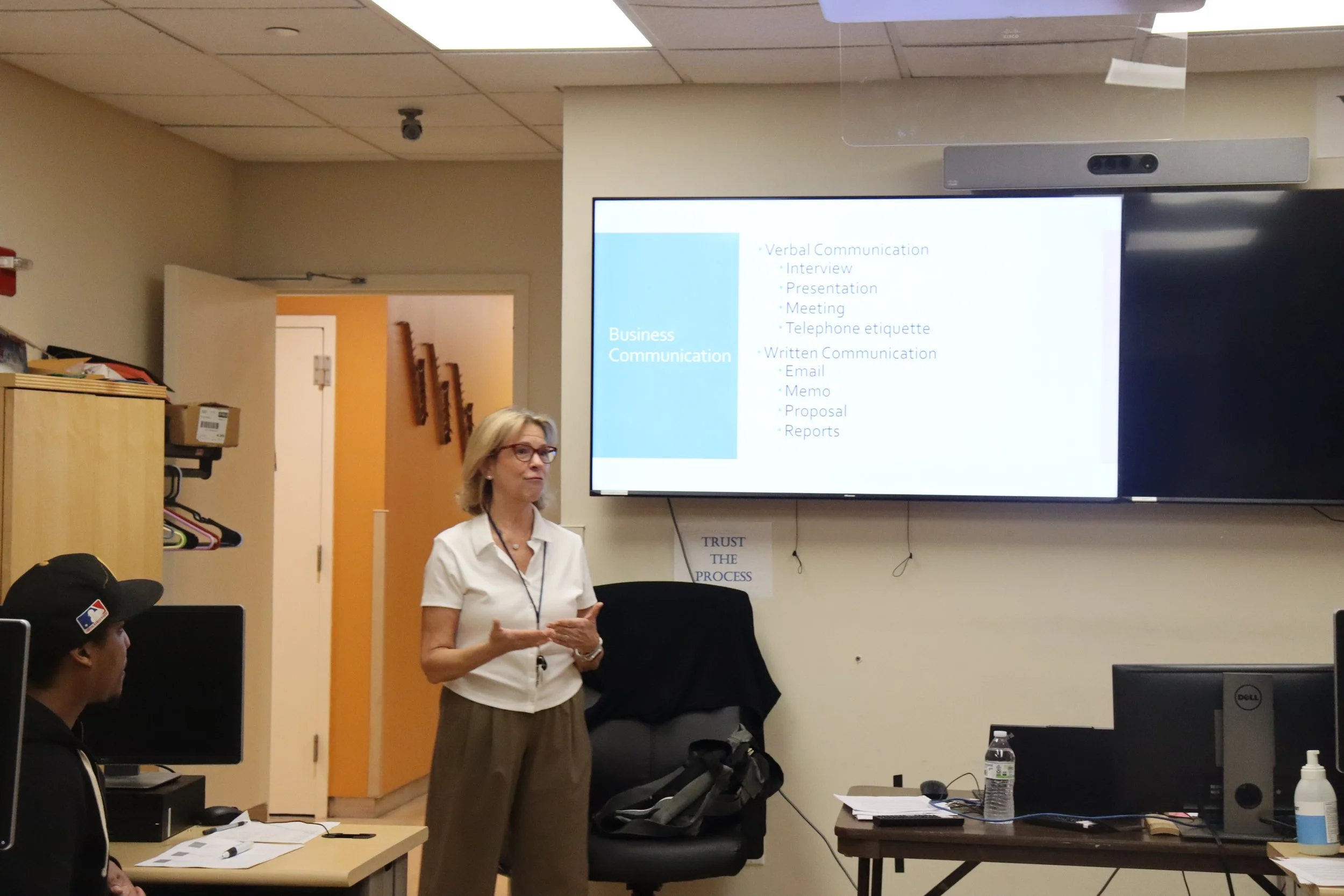 Woman giving a presentation in a classroom with a digital screen displaying business communication topics.