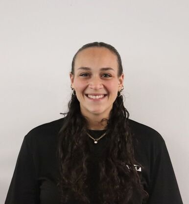 A young woman with dark curly hair, wearing a black shirt and jewelry, smiling in front of a plain white wall.
