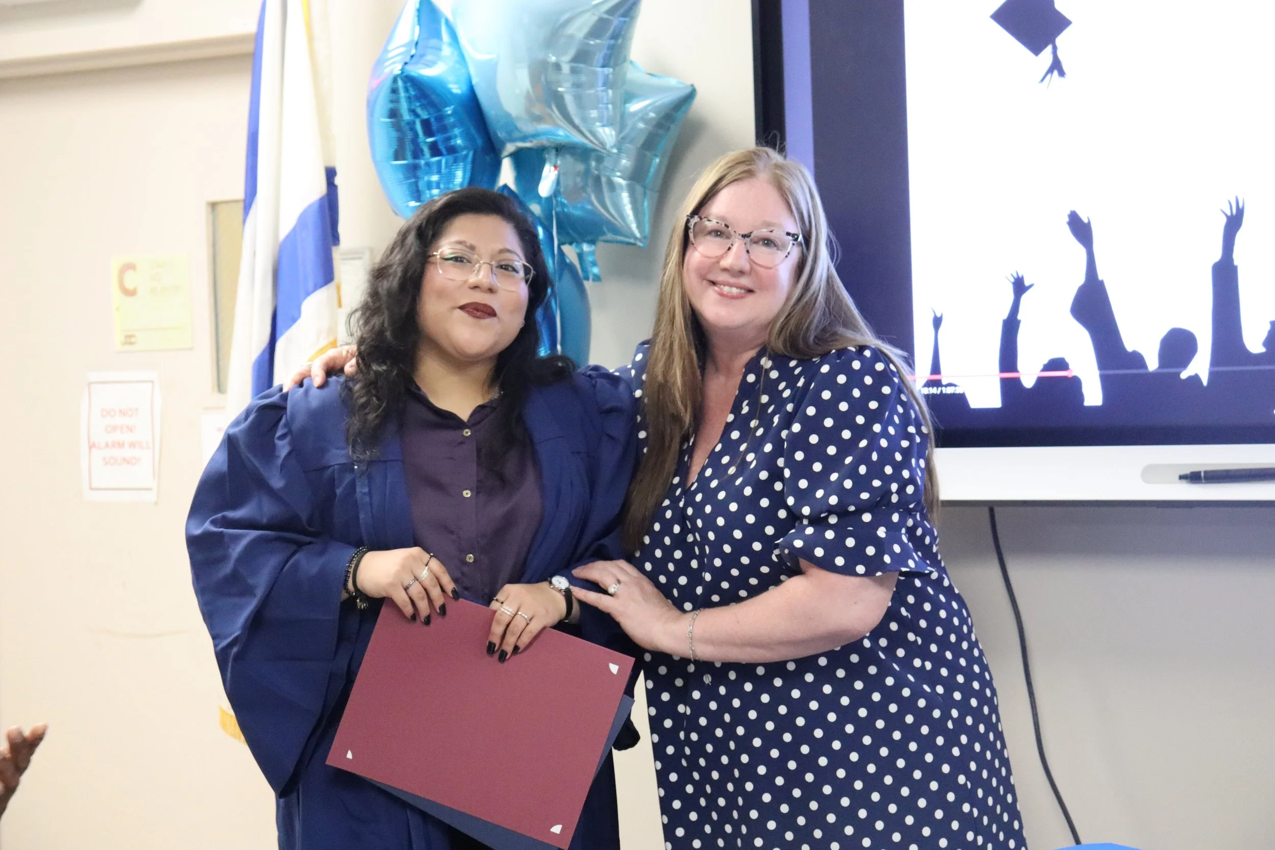 Two women standing close together, smiling, with one woman holding a large maroon-colored folder or certificate, in an indoor setting. Behind them, a screen displays a black silhouette of hands reaching up, and there are shiny blue balloons hanging on the wall.
