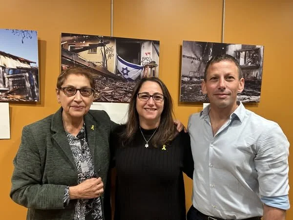 Three people standing together in front of a yellow wall with black and white photographs displayed behind them. The woman on the left is wearing glasses and a green blazer, the woman in the middle is smiling and wearing glasses with a black top, and the man on the right is dressed in a light blue shirt. They all have yellow ribbons pinned to their clothing.