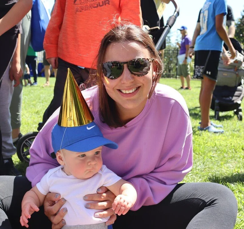 A young woman wearing sunglasses and a pink sweatshirt smiling while holding a baby in a blue Nike cap and white shirt at an outdoor gathering. The baby is wearing a gold party hat and a blue visor, looking off to the side. Several people are in the background on a grassy area.