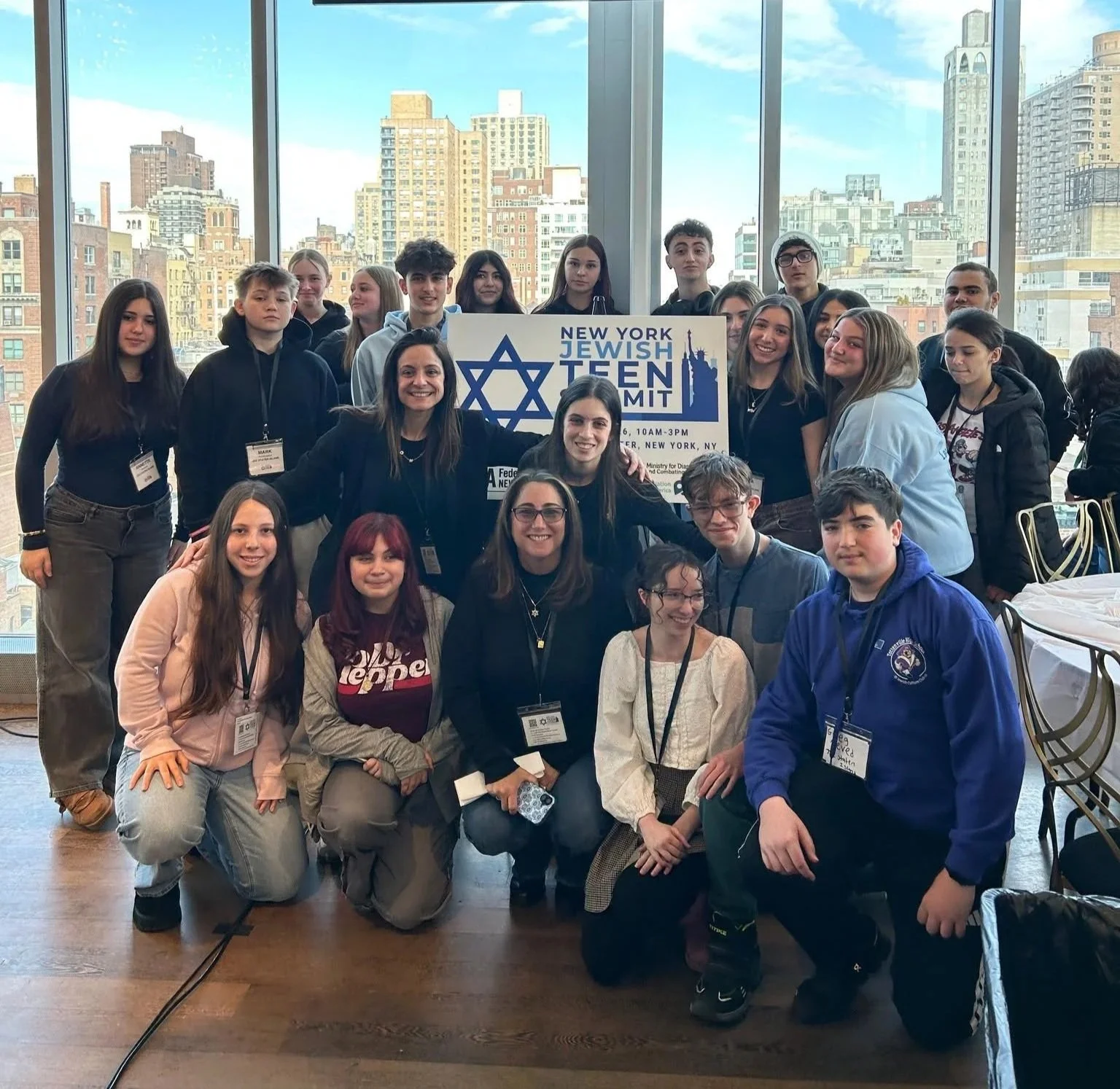 A group of young people and adults gathered indoors for a photo at the New York Jewish Teen Summit. They are standing in front of large windows overlooking New York City skyscrapers, with a sign displaying the event name and details. The group appears happy, with some smiling and posing for the camera.
