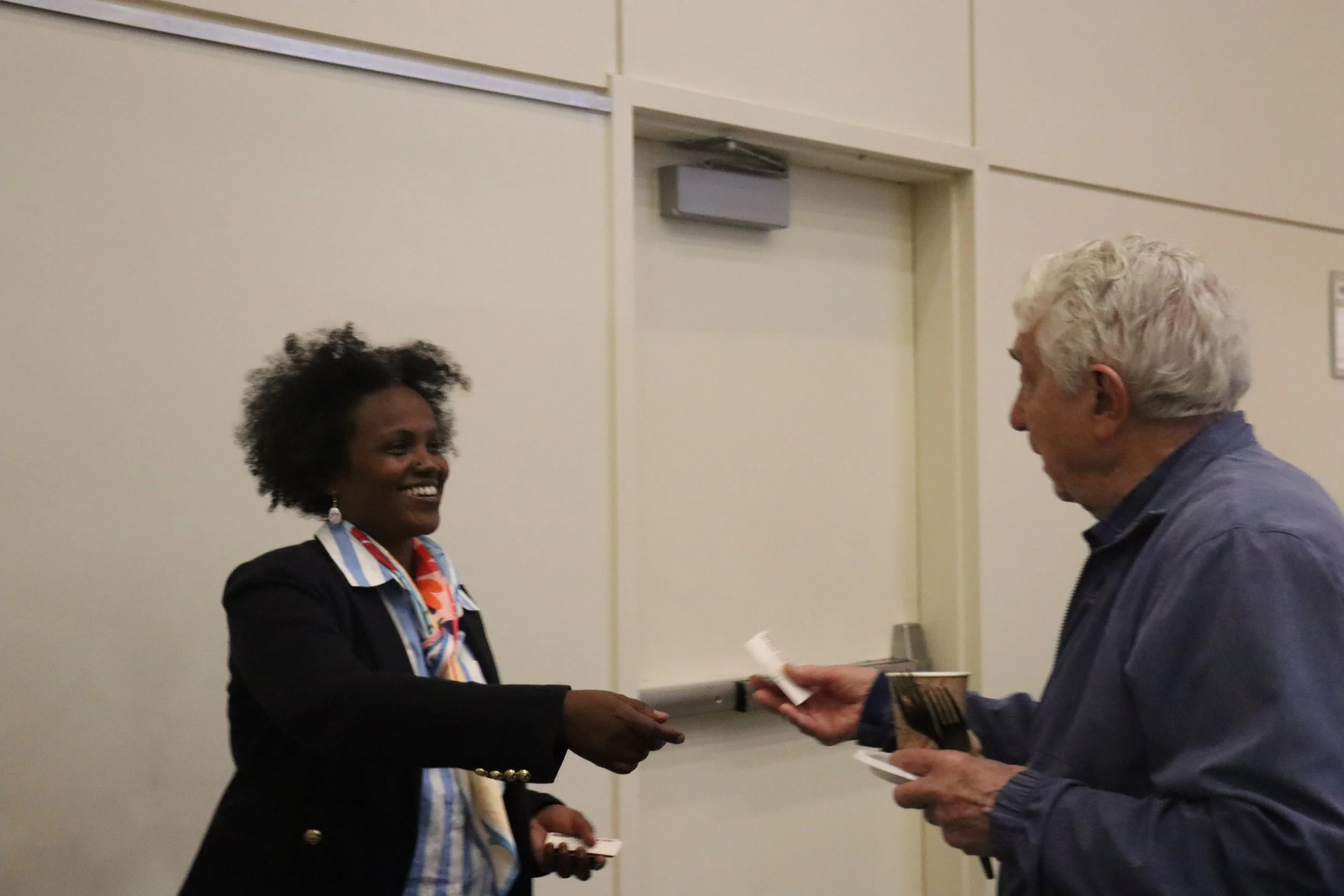 A woman in a business suit smiling as she receives a certificate or award from an older man with white hair, in an indoor setting with plain wall background.