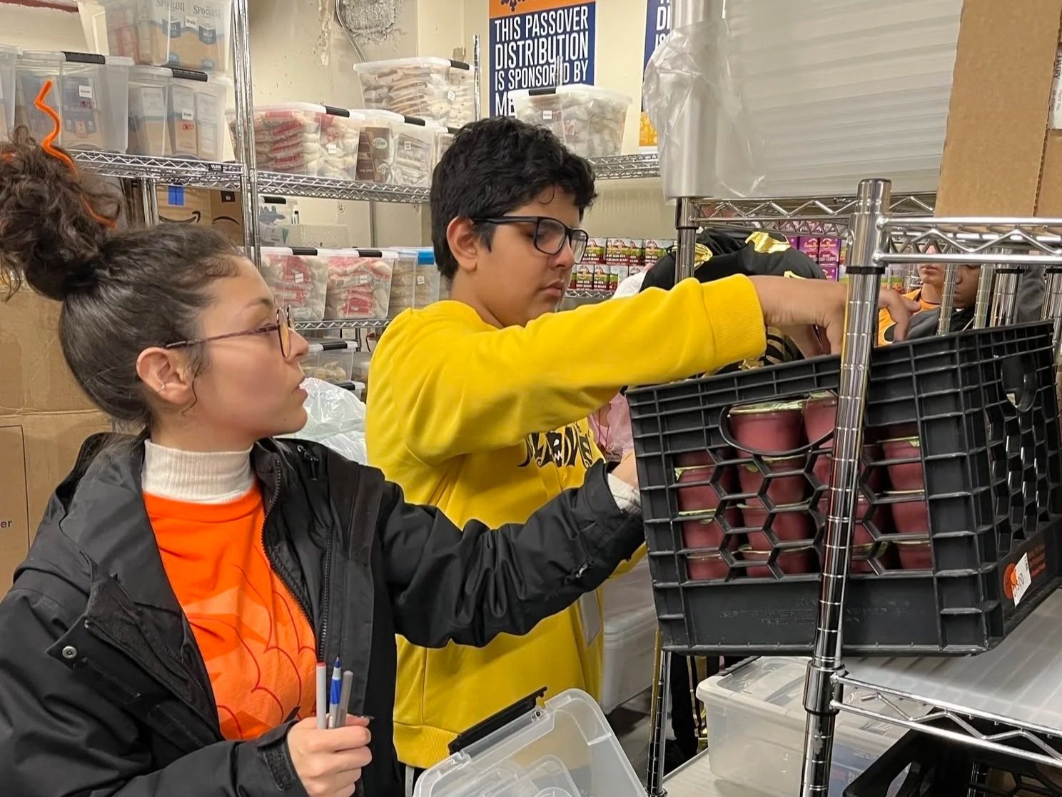 Two young people shopping for canned goods in a storage room. The girl with glasses, curly hair in a bun, and a black jacket holds pens. The boy with glasses and a yellow sweatshirt reaches into a black crate filled with canned goods on a metal shelving unit. Background features shelves with plastic containers and signs indicating support for a Passover distribution.