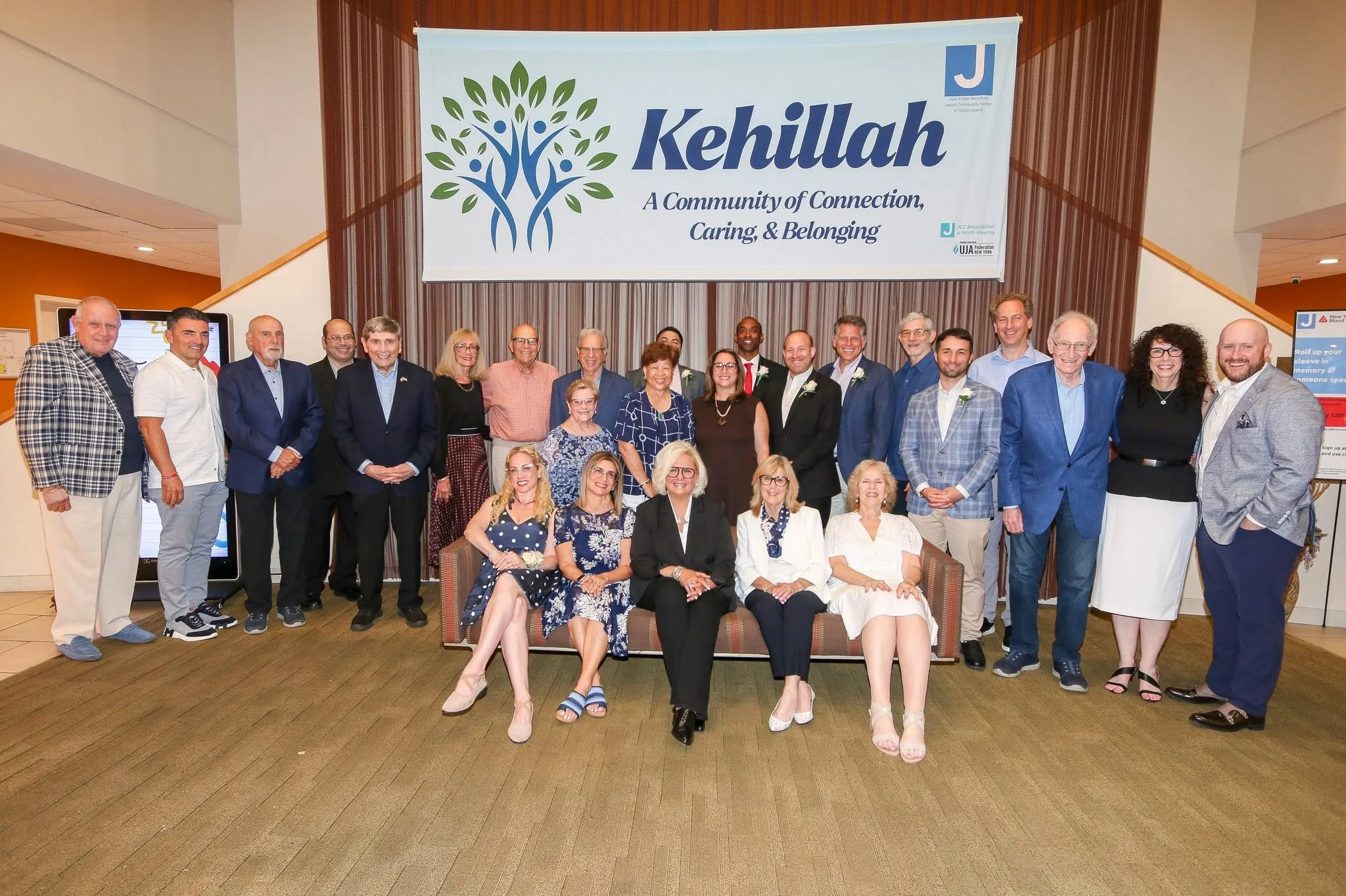 A group of people posing for a photo in front of a banner that reads 'Kehillah: A Community of Connection, Caring, & Belonging.' The group includes men and women of various ages, some dressed in suits and others in casual attire, standing and seated in a room with warm lighting and a wooden-paneled background.