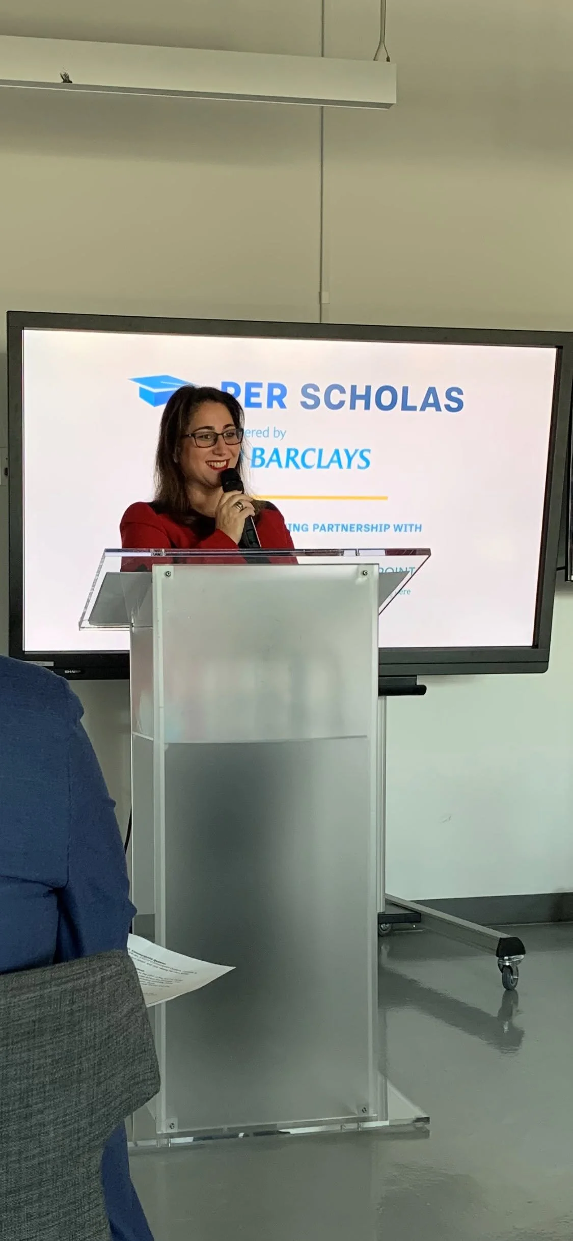 A woman in glasses and a red blazer is speaking into a microphone at a podium in front of a large screen displaying a presentation slide with the title 'PER SCHOLAS' and a logo of a graduation cap.