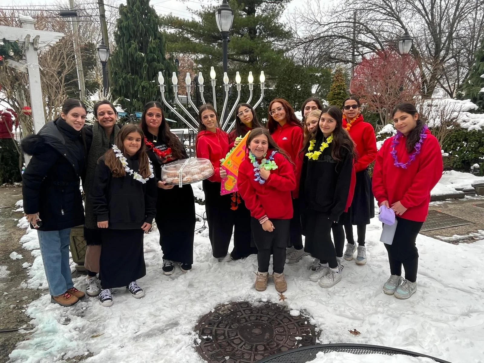 A group of women and girls standing outdoors in a snowy area, with a large menorah in the background, some wearing leis and holding a cake and a colorful book, celebrating Hanukkah.