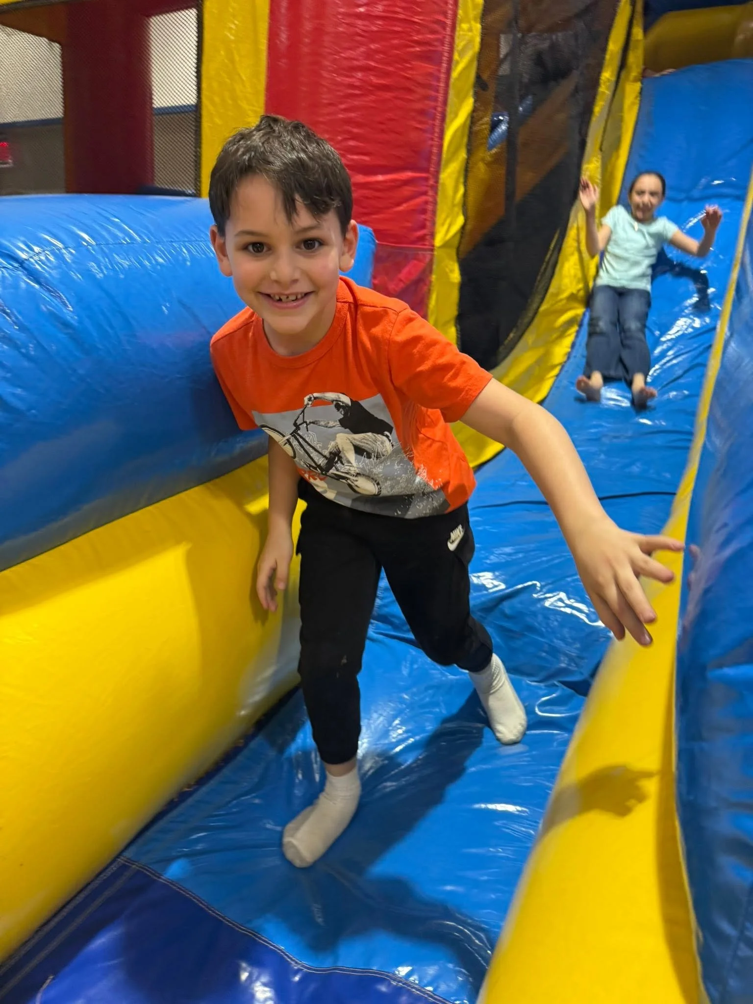 Children playing on an indoor inflatable bounce house with bright yellow, blue, red, and black colors.