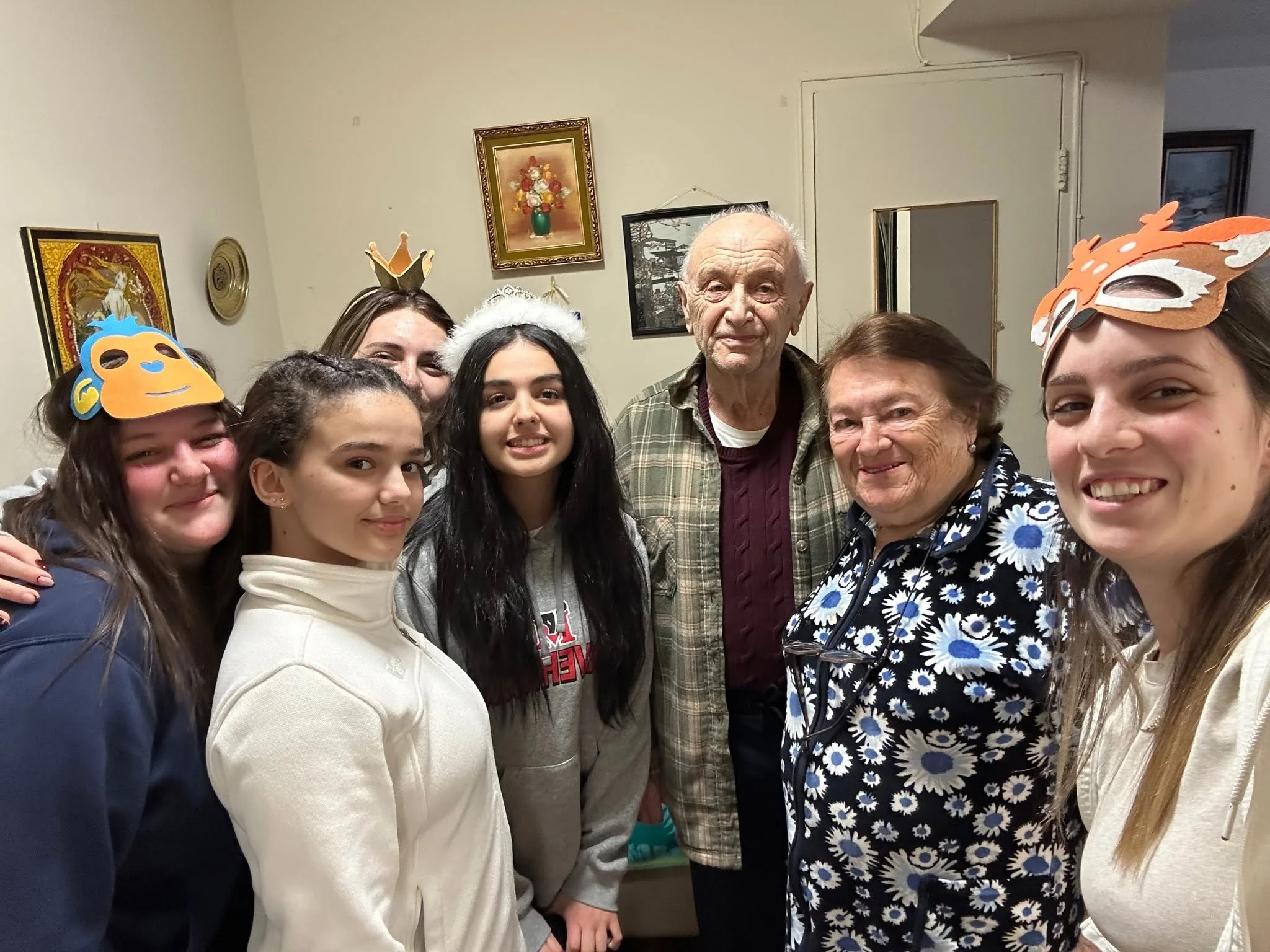 Group of young women and elderly individuals smiling and gathering together indoors, some wearing masks and headbands with decorative crowns, in a room with framed artwork on the walls.